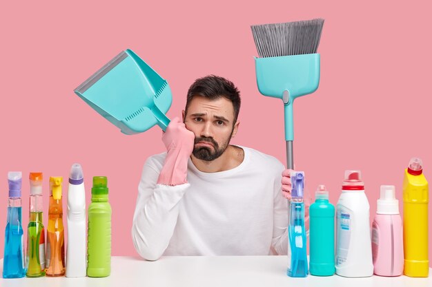 Upset tired husband wears pink rubber gloves, carries broom and scoop, takes break after sweeping floor, cleans house with cleanser