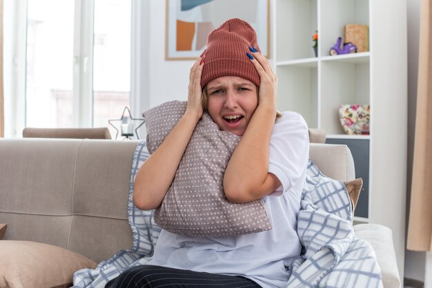 Upset unhealthy young woman in warm hat with blanket looking unwell and sick suffering from cold and flu holding pillow looking worried touching head sitting on couch in light living room