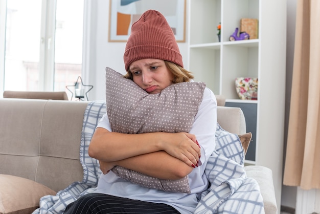 Upset unhealthy young woman in warm hat with blanket looking unwell and sick suffering from cold and flu holding pillow with sad expression on face sitting on couch in light living room