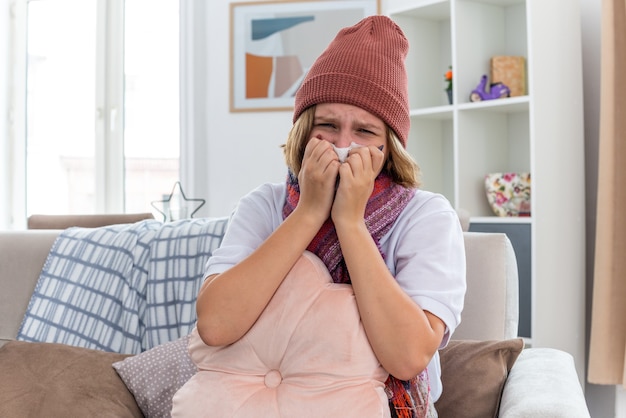 Upset unhealthy young woman in warm hat with scarf looking unwell and sick holding tissue suffering from cold and flu and blowing nose sneezing sitting on couch in light living room