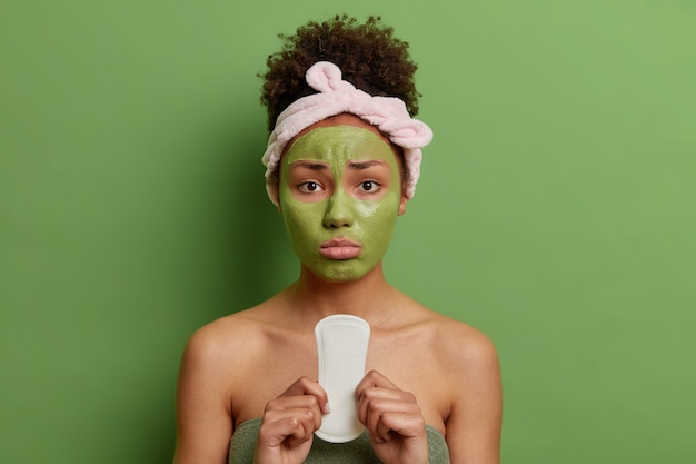 Free Photo upset woman with curly hair applies nourishing beauty mask on face holds sanitary napkin has mestruation suffers from pain wrapped in towel isolated over green wall