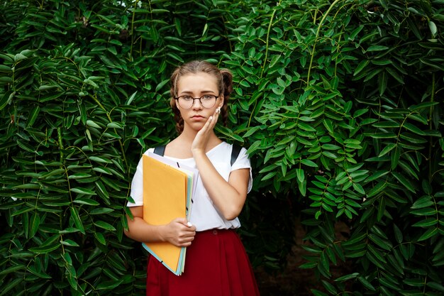 Upset young beautiful female student in glasses holding folders outdoors.