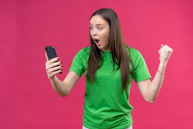 Upset young beautiful girl wearing green t-shirt holding smartphone looking at screen amazed and surprised standing over isolated pink background