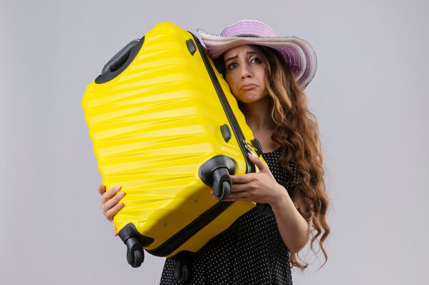 Upset young beautiful traveler girl in dress in polka dot in summer hat holding suitcase looking at camera with sad expression on face standing over white background