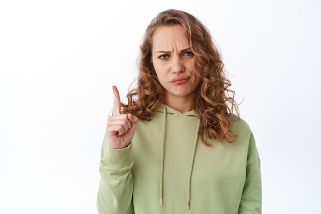 Free Photo upset young blond girl, frowning and looking disappointed, pointing finger up at top logo, judging bad thing, standing in green hoodie against white wall