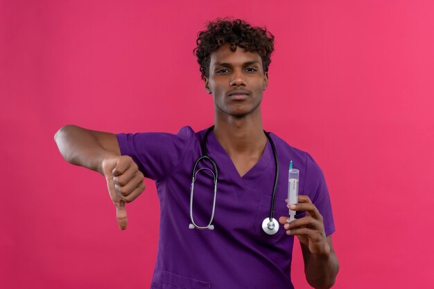 An upset young handsome dark-skinned doctor with curly hair wearing violet uniform with stethoscope showing thumbs down while holding plastic injection syringe 