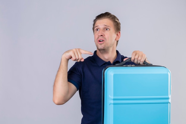 Upset young handsome traveler man holding suitcase looking away pointing finger to his suitcase standing over white background