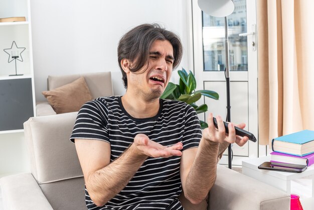 Upset young man in casual clothes holding tv remote crying hard sitting on the chair in light living room