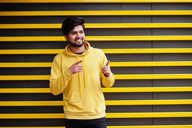 Free photo urban young hipster indian man in a fashionable yellow sweatshirt cool south asian guy wear hoodie against striped background showing fingers to free space