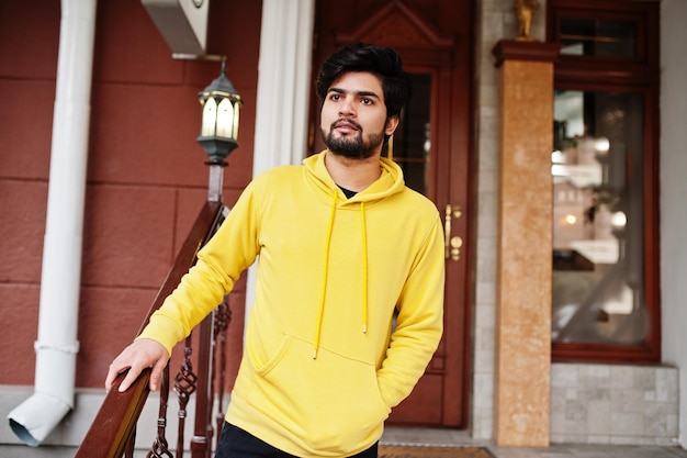 Free photo urban young hipster indian man in a fashionable yellow sweatshirt cool south asian guy wear hoodie stand on porch of the house