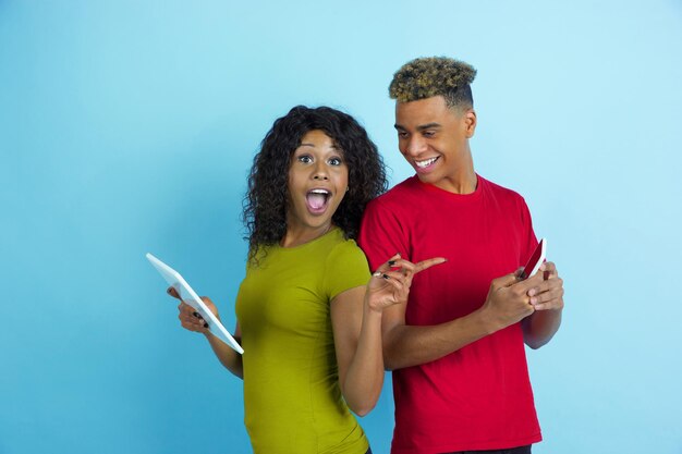 Using gadgets, laughting, pointing. Young emotional african-american man and woman in colorful clothes on blue background.