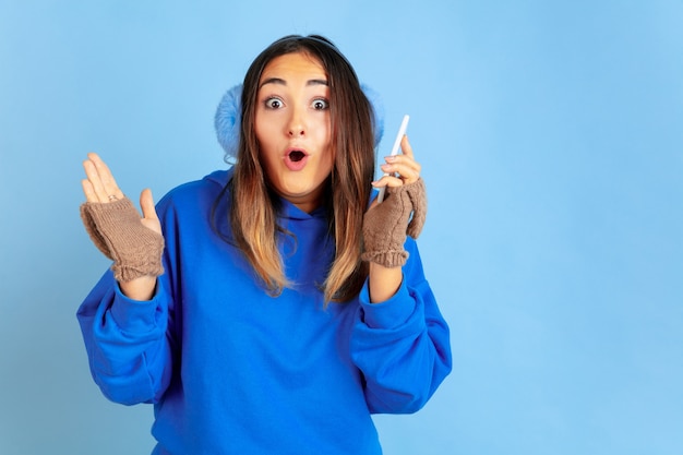 Using phone, happy. Caucasian woman's portrait on blue studio background. Beautiful female model in warm clothes. Concept of human emotions, facial expression, sales, ad. Winter mood, holidays.