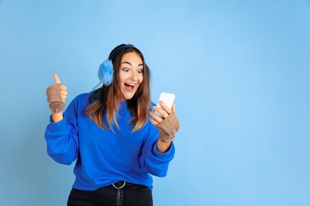 Using phone, happy. Caucasian woman's portrait on blue studio background. Beautiful female model in warm clothes. Concept of human emotions, facial expression, sales, ad. Winter mood, holidays.