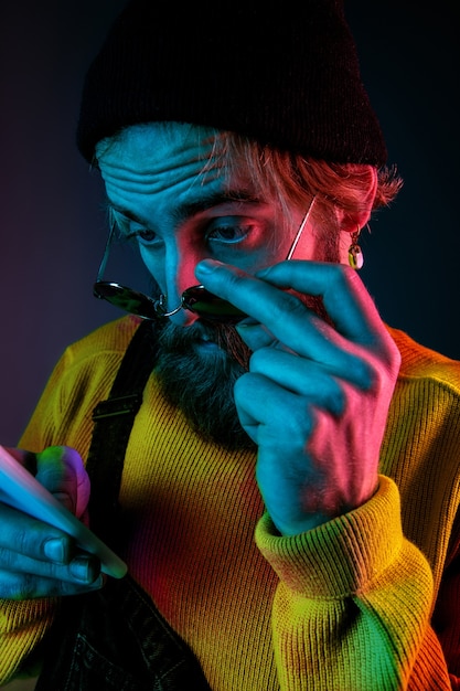 Using phone in sunglasses. Caucasian man's portrait on gradient studio background in neon light. Beautiful male model with hipster style. Concept of human emotions, facial expression, sales, ad.