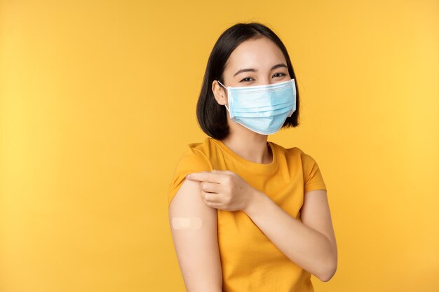 Vaccination and covid19 pandemic concept Smiling asian woman in medical face mask showing her shoulder with band aid after vaccinating from coronavirus yellow background