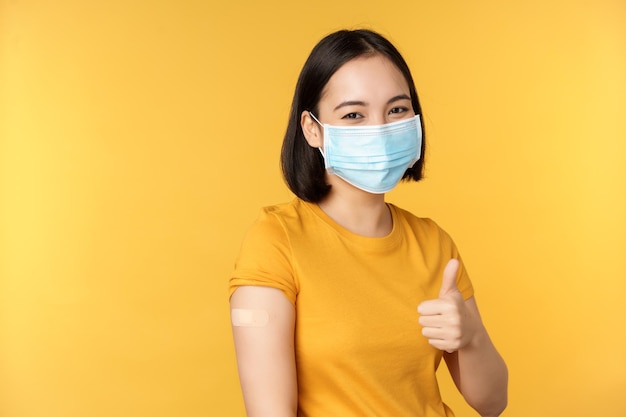 Vaccination from covid and health concept Happy asian girl showing thumbs up wearing medical mask band aid on shoulder got coronavirus vaccine shot yellow background