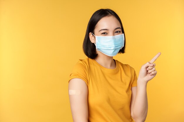 Vaccination from covid and health concept Image of smiling korean girl in medical face mask band aid on shoulder pointing finger at banner advertisement yellow background