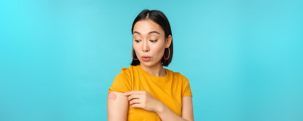 Vaccine campaign from covid19 Young beautiful healthy asian woman showing shoulder with bandaid concept of vaccination standing over blue background