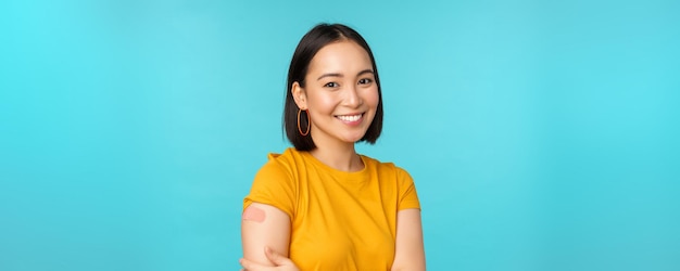 Vaccine campaign from covid19 Young beautiful healthy asian woman showing shoulder with bandaid concept of vaccination standing over blue background