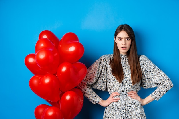 Free Photo valentines day. angry and confused girlfriend in dress, standing near red heart balloons and frowning annoyed at camera, standing near blue background.