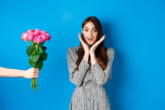 Valentines day concept. Excited and happy young woman looking amazed at camera while hand stretch out hand with bouquet of flowers, receiving romantic gift, blue background