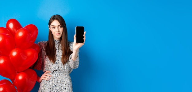 Free photo valentines day pretty smiling woman in dress showing empty smartphone screen standing near romantic