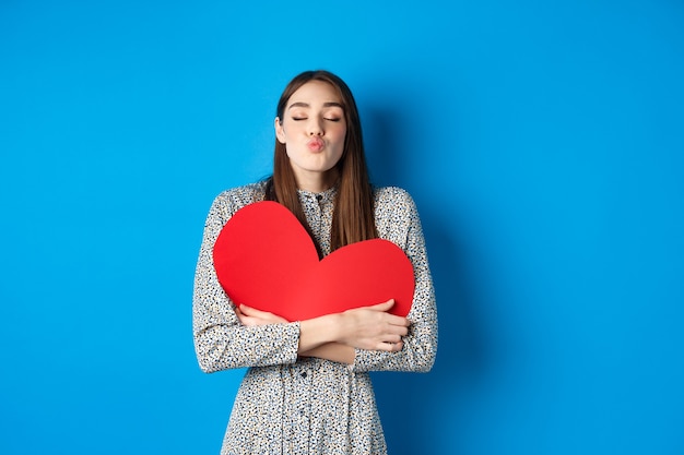 Valentines day romantic beautiful woman close eyes and pucker lips for kiss holding big red heart cu...