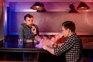 Free photo vaping man holding a mod. a cloud of vapor at the vapebar. two men rest in a bar and smoke electronic cigarettes.