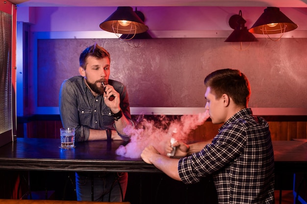 Vaping man holding a mod. A cloud of vapor at the vapebar. Two men rest in a bar and smoke electronic cigarettes.