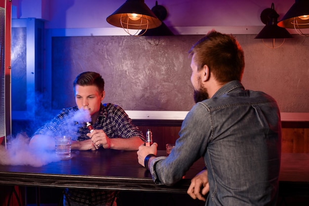 Vaping man holding a mod. A cloud of vapor at the vapebar. Two men rest in a bar and smoke electronic cigarettes.