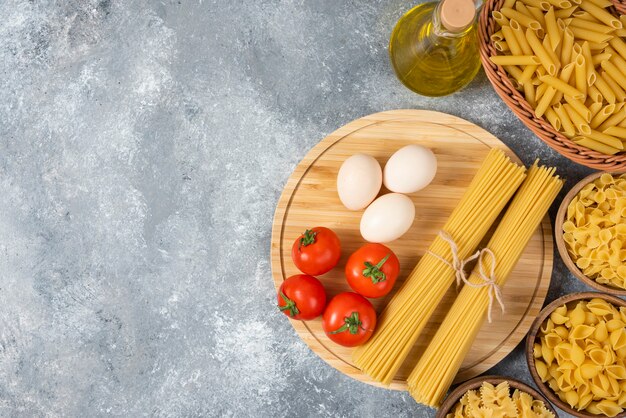 Variety of raw pasta with eggs, fresh tomatoes and bottle of oil on marble surface.