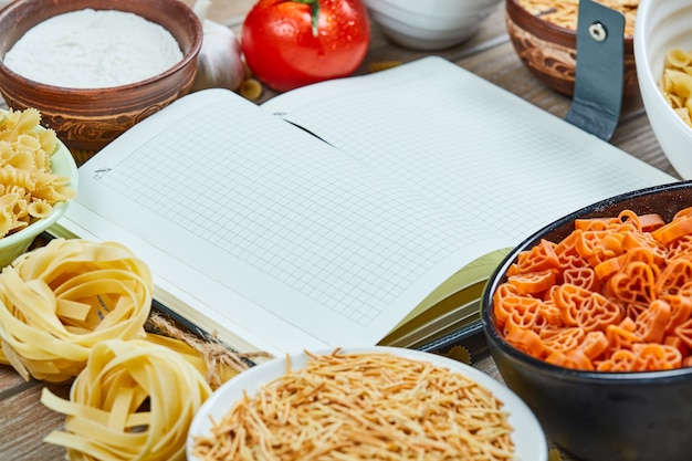 Free photo various uncooked pasta with notebook and vegetables on a wooden table.