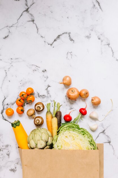 Vegetables spill out from brown paper bag on marble background