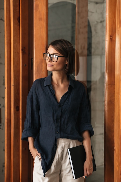 Vertical image of business woman in eyeglases with folder in hand standing near the glass door