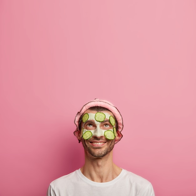 Free photo vertical image of handsome happy man applies slices of cucumber, wears bath hat, enjoys beauty treatments