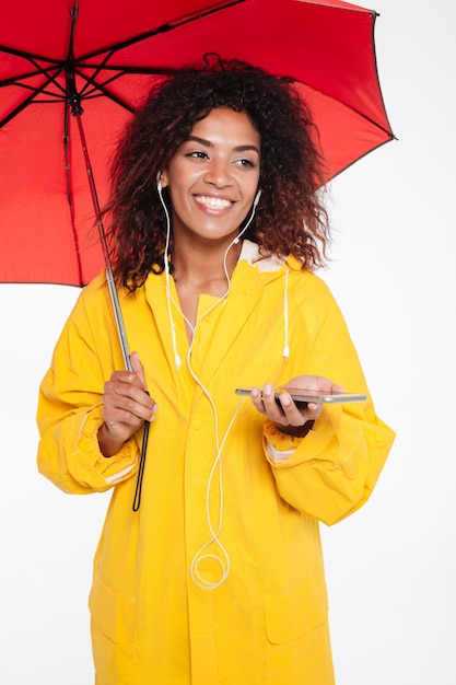 Vertical image of happy african woman in raincoat hiding under umbrella and listening music on her phone while looking away over white