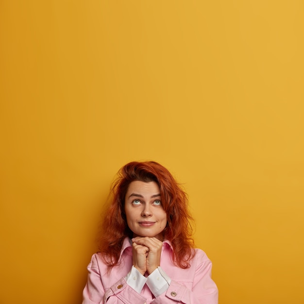 Free photo vertical image of pensive lovely woman looks hopefully above, keeps hands under chin and prays for good wellness, has red hair