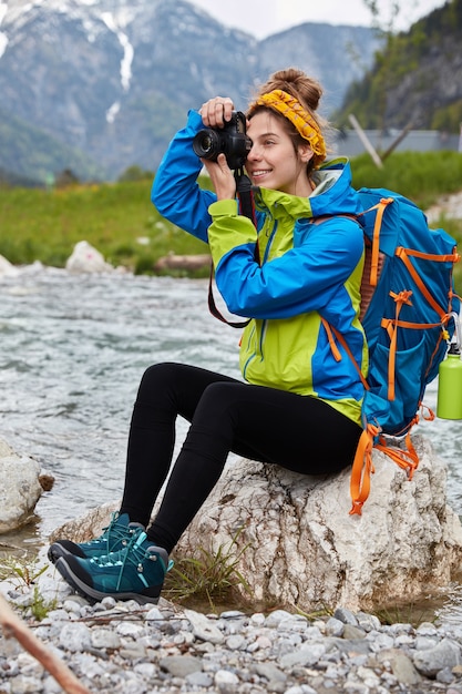 Vertical outdoor shot of cheerful female makes professional photos, sits on rocks near mountain river