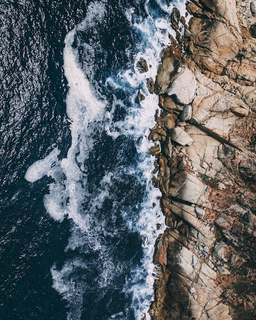 Free photo vertical overhead shot of rocky shoreline next to a body of water with waves splashing the rocks