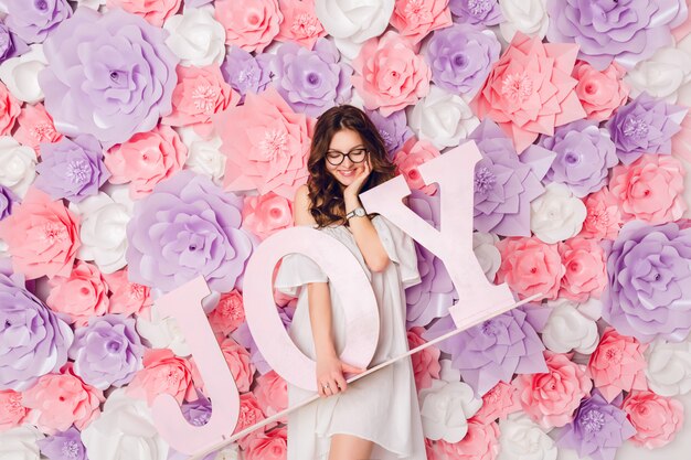 Vertical portrait of cute brunette girl. She stands and holds wood word JOY smiling widely. Background in pink flowers