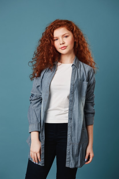 Vertical portrait of good-looking teen woman with wavy ginger hair and freckles in white t-shirt and grey cardigan posing for school graduation album with happy expression.