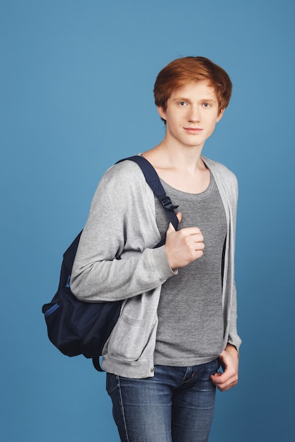 Vertical portrait of serious young red-headed male student in casual gray outfit with black backpack, holding hand in pocket,  with relaxed and confident expression