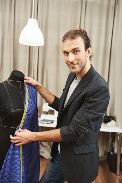 Vertical portrait of young attractive professional caucasian male designer in stylish outfit working on new dress collection for following fashion show, checking neckline size using measuring tape