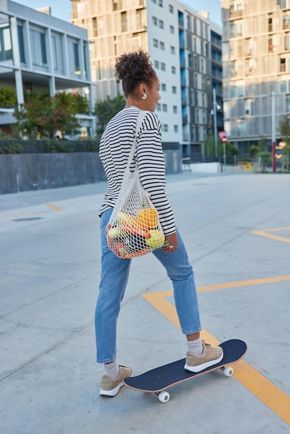 Vertical shot of active curly haired woman rides skateboard poses back to camera spends free time actively outdoors dressed in street clothes carries bag poses in city during daytime sunny weather