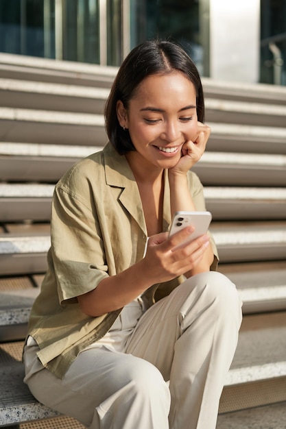 Vertical shot of asian woman student sits on stairs in city looking at mobile phone screen and smili