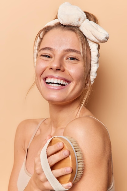 Free photo vertical shot of attractive young european woman smiles happily use body brush for skin care enjoys spa treatment wears headband casual t shirt isolated over beige background. scrubbing and message