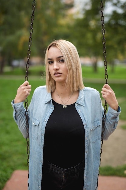 Vertical shot of a beautiful Caucasian woman in denim holding a metal swing in the park