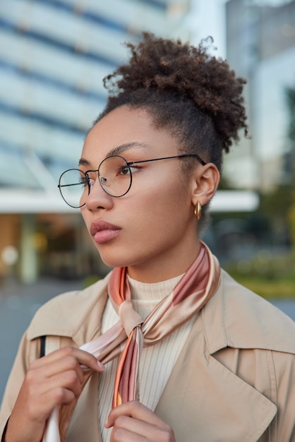 Free photo vertical shot of beautiful curly haired millennial gir wears round spectacles for vision correction coat and tied kerchief around neck looks thoughtfully forward while strolling outside during daytime