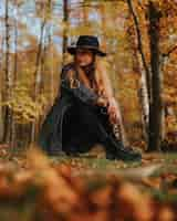 Free photo vertical shot of a blonde female model with black coat and hat posing in an autumnal forest