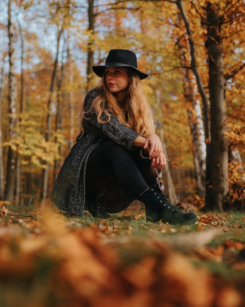 Free Photo vertical shot of a blonde female model with black coat and hat posing in an autumnal forest